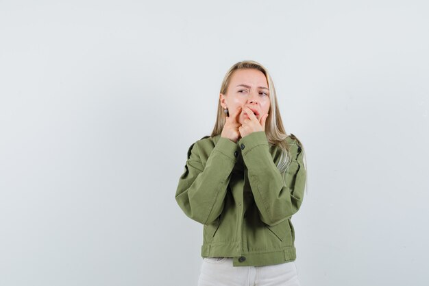 Giovane donna che stringe il suo brufolo sulla guancia in giacca, pantaloni, vista frontale.