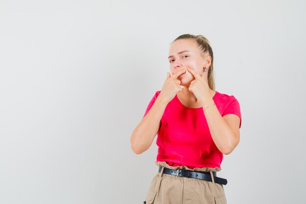 Giovane donna che stringe il suo brufolo sulla guancia in t-shirt, pantaloni, vista frontale.