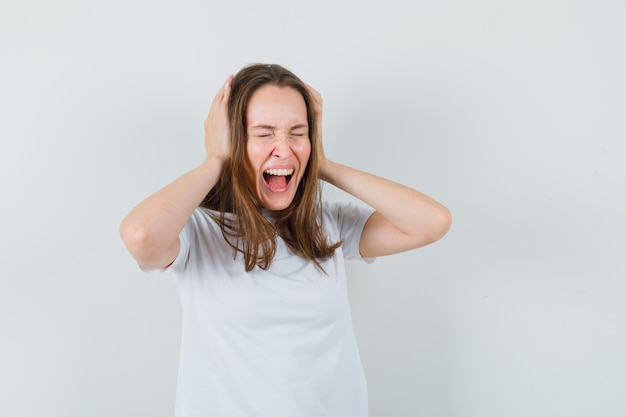 Giovane donna che stringe la testa mentre urla in maglietta bianca e sembra eccitata