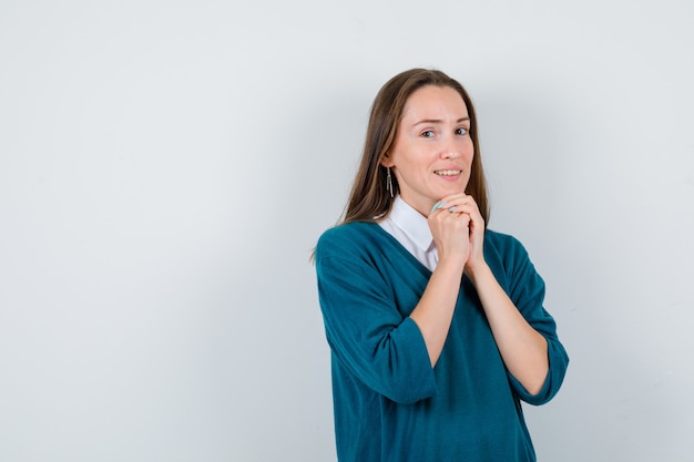 Giovane donna che stringe le mani nel gesto di preghiera in maglione sopra la camicia bianca e sembra speranzosa, vista frontale.