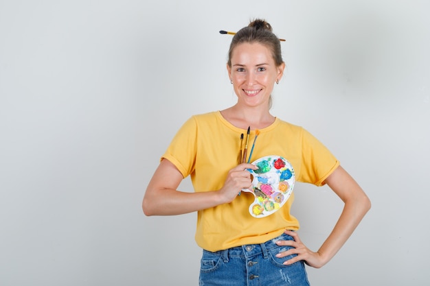 Giovane donna che tiene gli strumenti di pittura con la mano sulla vita in maglietta gialla, pantaloncini di jeans e sembra allegro