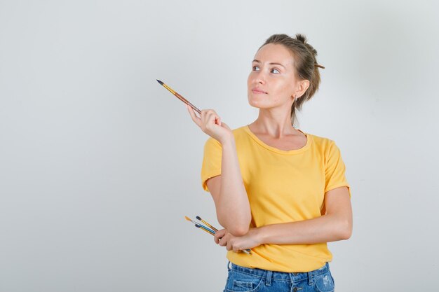 Giovane donna che tiene gli strumenti di pittura e distoglie lo sguardo in maglietta gialla