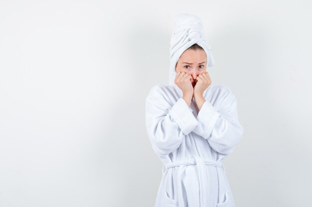 Giovane donna che tiene i pugni sulle guance in accappatoio bianco, asciugamano e sembra spaventata, vista frontale.