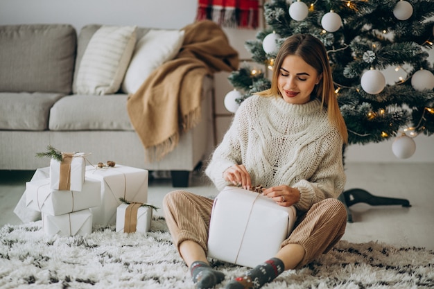 Giovane donna che tiene i regali di Natale e che si siede sotto l'albero di Natale