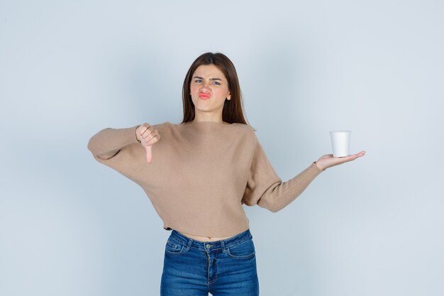 Giovane donna che tiene il bicchiere di carta, mostrando il pollice in giù in maglione, jeans e guardando dispiaciuto, vista frontale.