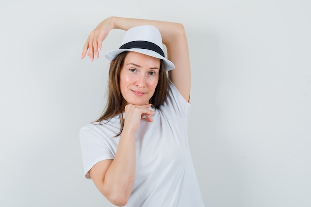 Giovane donna che tiene il braccio alzato sulla testa in cappello bianco della maglietta e sembra ragionevole