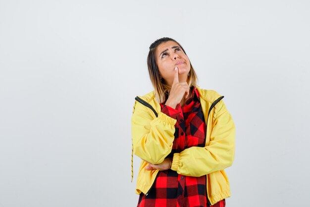 Giovane donna che tiene il dito sul mento, alzando lo sguardo in camicia a scacchi, giacca e guardando pensieroso, vista frontale.