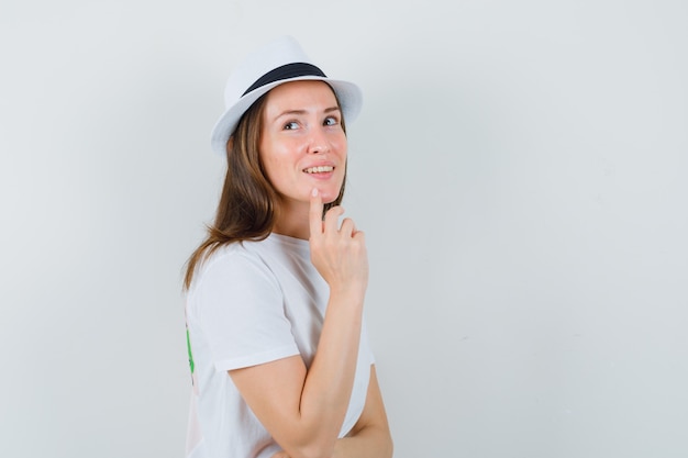 Giovane donna che tiene il dito sul mento in maglietta bianca, cappello e sguardo sognante.