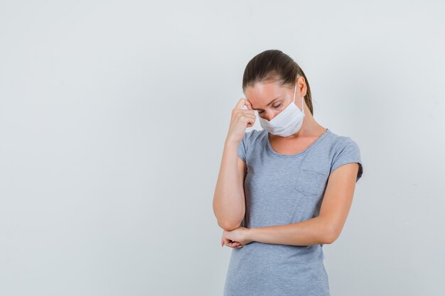 Giovane donna che tiene il dito sulla fronte in maglietta grigia, maschera e sembra esausta. vista frontale.