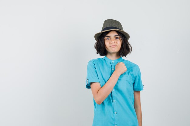 Giovane donna che tiene il pugno sul petto in camicia blu, cappello e aspetto maestoso.