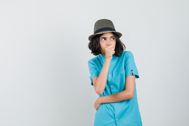 Giovane donna che tiene il pugno sulla sua bocca mentre distoglie lo sguardo in camicia blu, cappello e sembra ansioso.