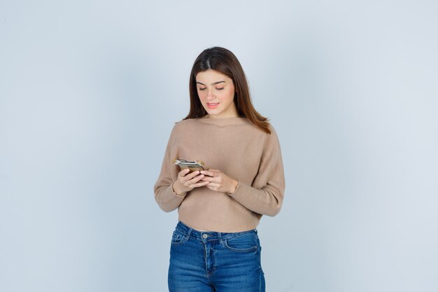 Giovane donna che tiene il telefono cellulare in maglione beige, jeans e sembra soddisfatta. vista frontale.