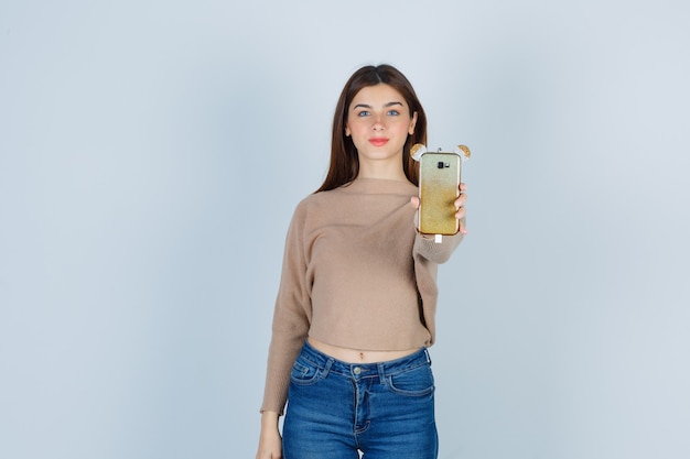 Giovane donna che tiene il telefono cellulare in maglione beige, jeans e sembra soddisfatta. vista frontale.