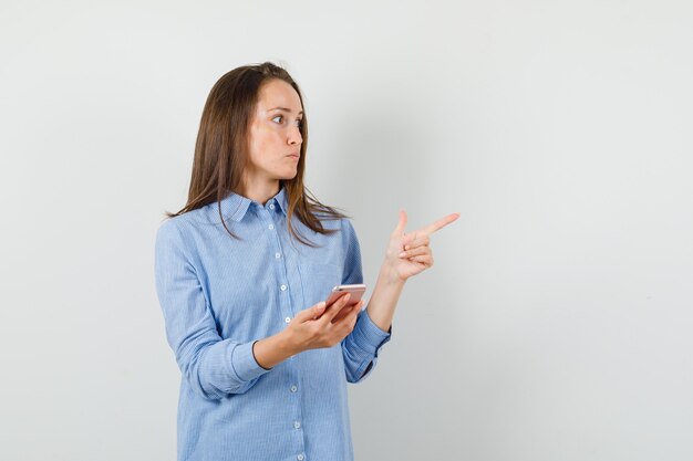 Giovane donna che tiene il telefono cellulare mentre punta lontano in maglietta blu e guardando concentrato.