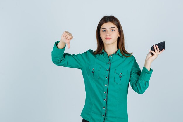 Giovane donna che tiene il telefono cellulare, mostrando il pollice verso il basso in camicia verde e guardando dispiaciuto, vista frontale.