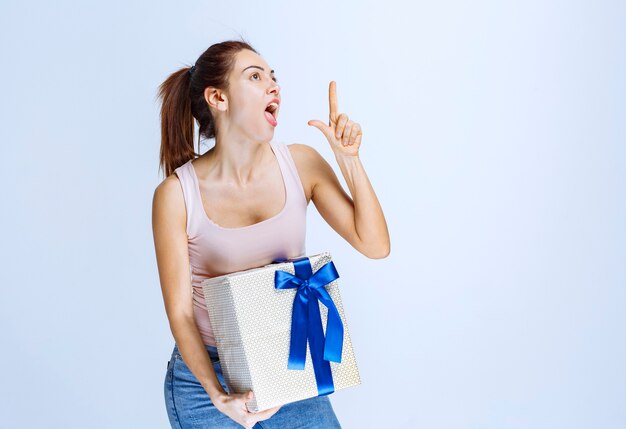 Giovane donna che tiene in mano una scatola regalo bianca avvolta con un nastro blu e lo dimostra