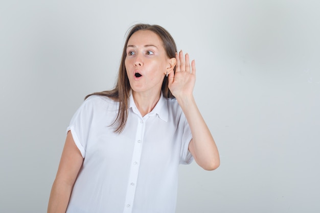 Giovane donna che tiene la mano all'orecchio in camicia bianca e che sembra sorpreso