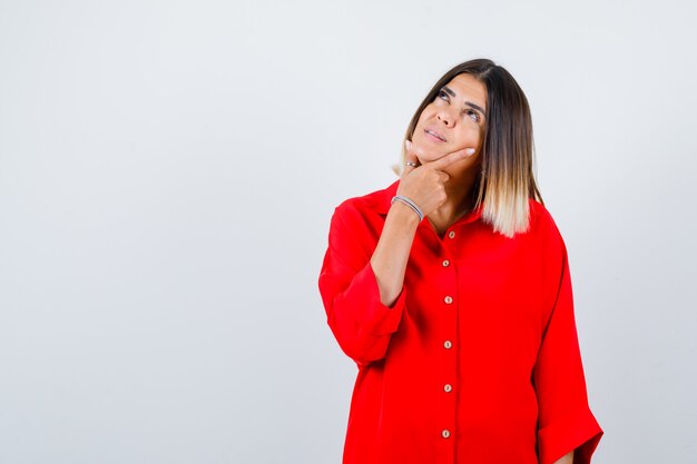 Giovane donna che tiene la mano sotto il mento in una camicia oversize rossa e guarda una vista frontale premurosa.