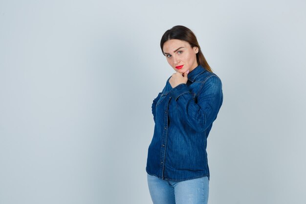 Giovane donna che tiene la mano sul fianco mantenendo la mano sul mento in camicia di jeans e jeans e sembra affascinante