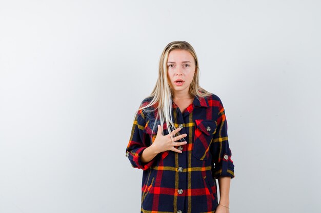 Giovane donna che tiene la mano sul petto in camicia a quadri e sembra perplessa, vista frontale.