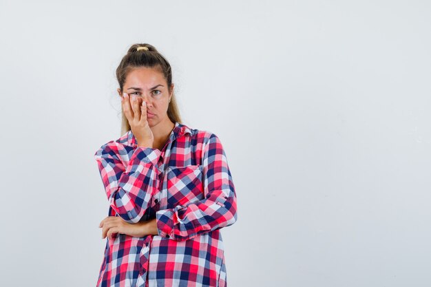 Giovane donna che tiene la mano sul viso in camicia a quadri e sembra triste, vista frontale.