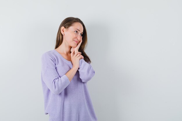 Giovane donna che tiene la mano sul viso mentre guarda da parte in camicetta lilla e sembra felice. vista frontale. spazio per il testo