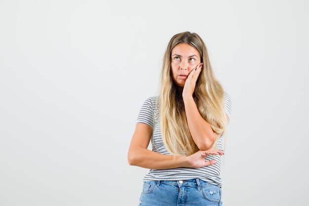 Giovane donna che tiene la mano sul viso mentre guarda lontano in t-shirt e guardando pensieroso, vista frontale.