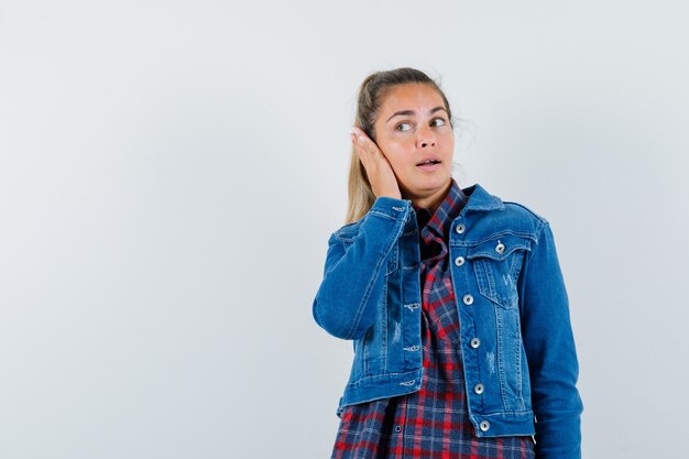 Giovane donna che tiene la mano sull'orecchio in camicia, giacca e sguardo curioso. vista frontale.