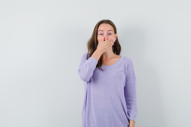 Giovane donna che tiene la mano sulla bocca in camicia casual e sembra allegra. vista frontale.