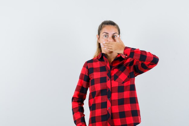 Giovane donna che tiene la mano sulla bocca in camicia casual e sembra spaventata. vista frontale.