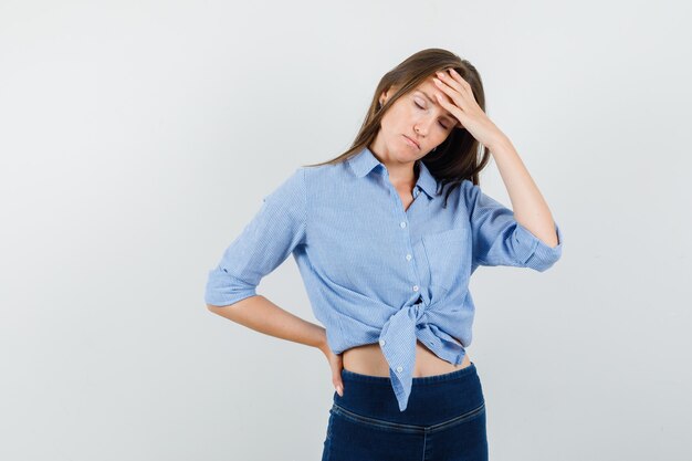 Giovane donna che tiene la mano sulla fronte in camicia blu, pantaloni e sembra affaticata.