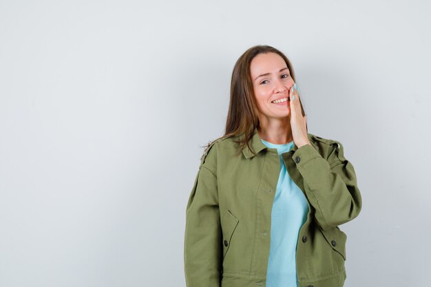 Giovane donna che tiene la mano sulla guancia in t-shirt, giacca e sembra carina, vista frontale.