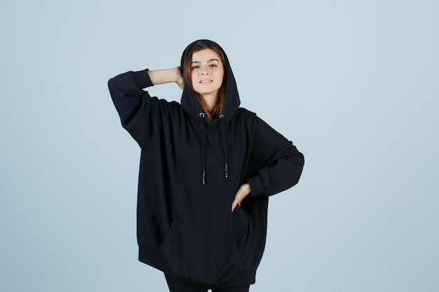 Giovane donna che tiene la mano sulla testa mentre tiene la mano sulla vita in felpa con cappuccio oversize, pantaloni e sembra fresca, vista frontale.