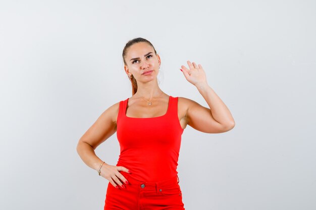 Giovane donna che tiene la mano sulla vita, mostrando il gesto di arresto in canottiera rossa, pantaloni e guardando fiducioso, vista frontale.