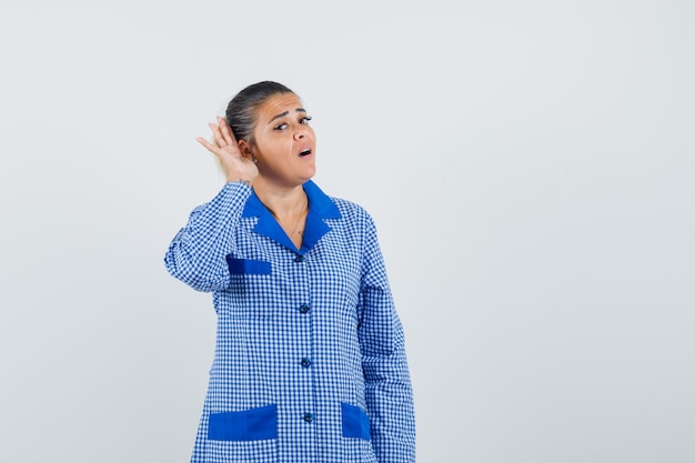 Giovane donna che tiene la mano vicino all'orecchio mentre ascolta qualcuno in camicia del pigiama a quadretti blu e sembra concentrata. vista frontale.