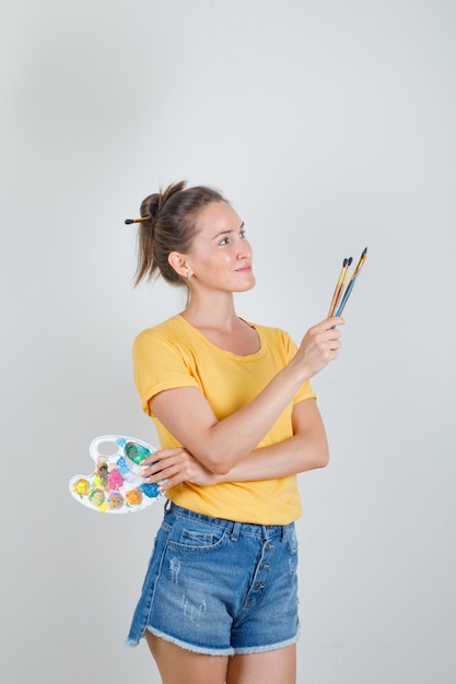 Giovane donna che tiene la tavolozza di arte con i pennelli in maglietta gialla, pantaloncini di jeans e sembra felice