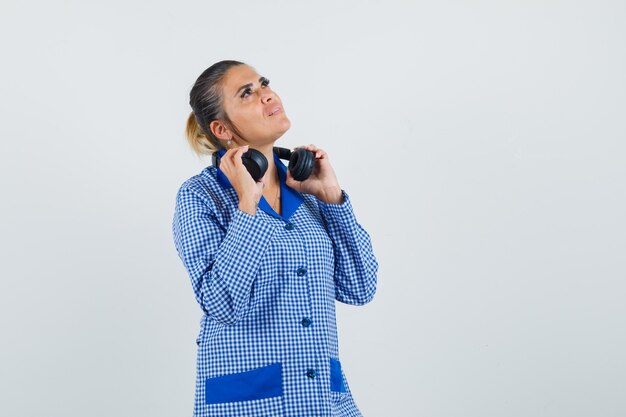 Giovane donna che tiene le cuffie e guardando sopra in camicia pigiama a quadretti blu e guardando pensieroso. vista frontale.