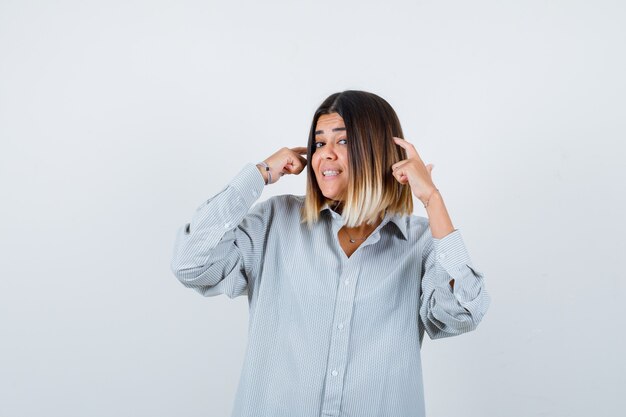 Giovane donna che tiene le dita sulla testa in camicia oversize e sembra allegra. vista frontale.