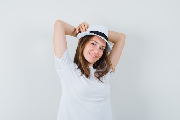 Giovane donna che tiene le mani alzate dietro la testa in maglietta bianca, cappello e sguardo rilassato.