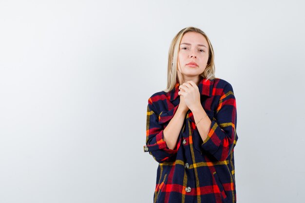 Giovane donna che tiene le mani giunte per pregare in camicia a quadri e guardando pacifica, vista frontale.