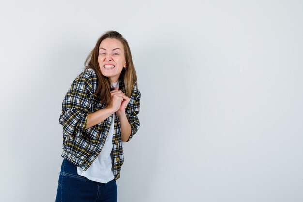 Giovane donna che tiene le mani giunte sul petto in t-shirt, giacca, jeans e che sembra felice, vista frontale.