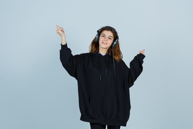 Giovane donna che tiene le mani in alto mentre ascolta la musica con handphones in felpa oversize, pantaloni e sembra energica vista frontale.