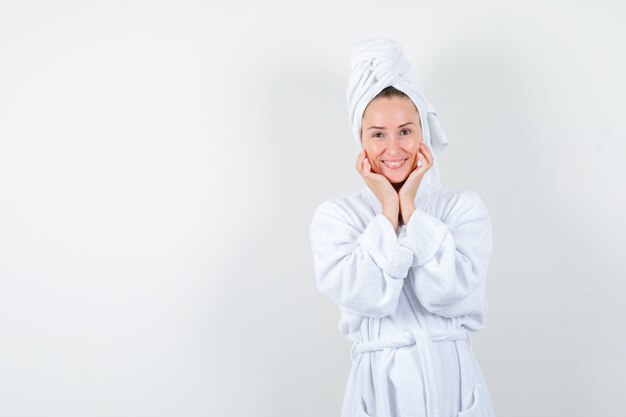Giovane donna che tiene le mani sotto il mento in accappatoio bianco, asciugamano e guardando allegro, vista frontale.