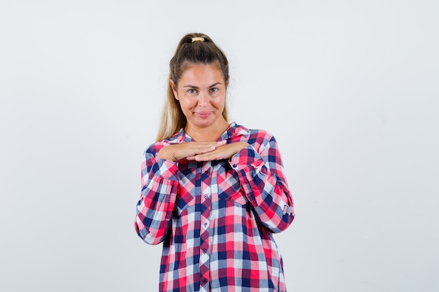 Giovane donna che tiene le mani sotto il mento in camicia a quadri e sembra adorabile. vista frontale.