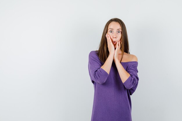 Giovane donna che tiene le mani sul viso in camicia viola e guardando scioccato, vista frontale.