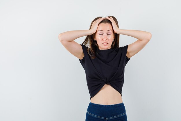 Giovane donna che tiene le mani sulla testa in camicetta nera, pantaloni e sembra affaticata.