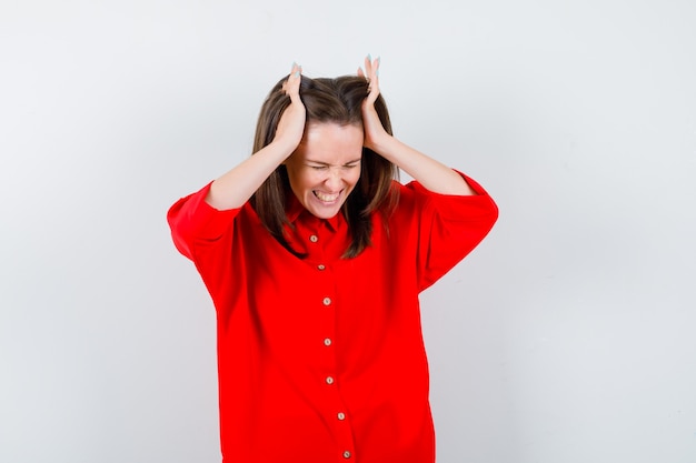Giovane donna che tiene le mani sulla testa in camicetta rossa e sembra irritata, vista frontale.