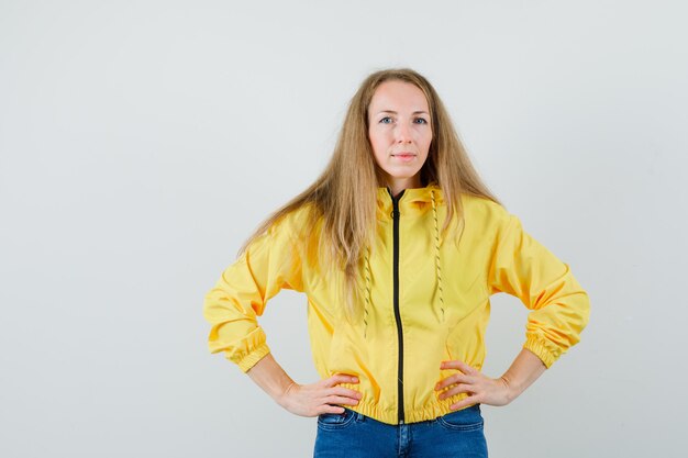 Giovane donna che tiene le mani sulla vita in bomber giallo e jeans blu e sembra attraente, vista frontale.