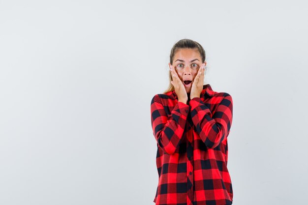 Giovane donna che tiene le mani sulle guance in camicia a quadri e sembra scioccata