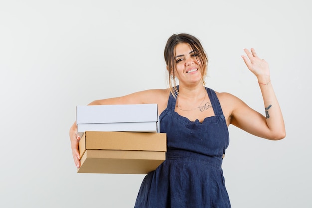 Giovane donna che tiene scatole di cartone, agitando la mano nel vestito e guardando allegra